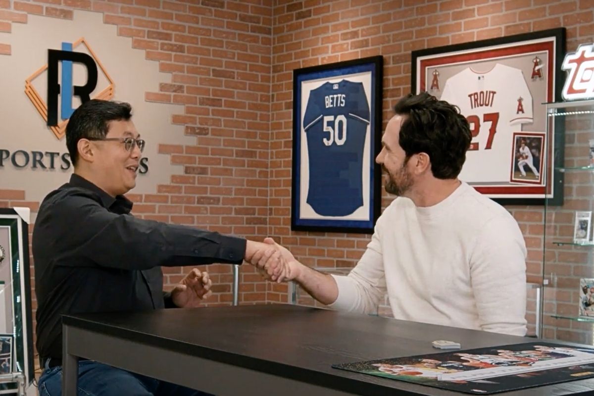 Two men shaking hands at a table with sports memorabilia in the background